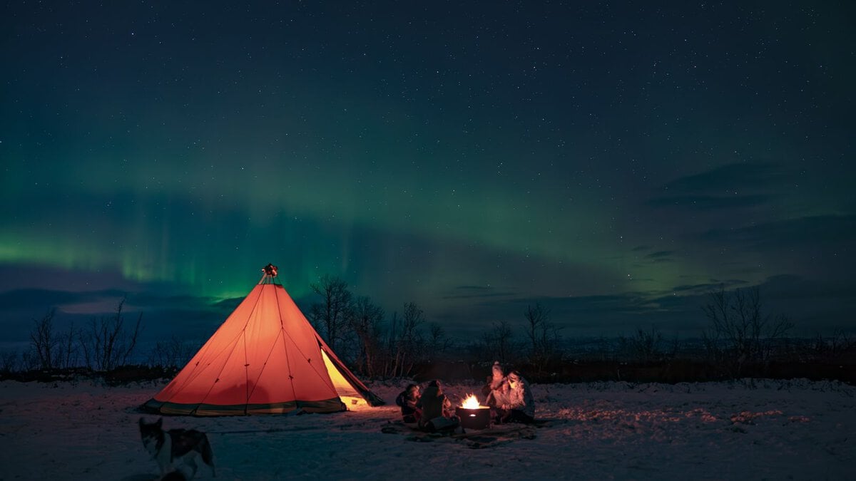 hiking with huskies in abisko - An Evening Walk With Huskies Northern Lights glowing above a traditional lávvu tent in the snowy Arctic landscape of Abisko, Swedish Lapland.